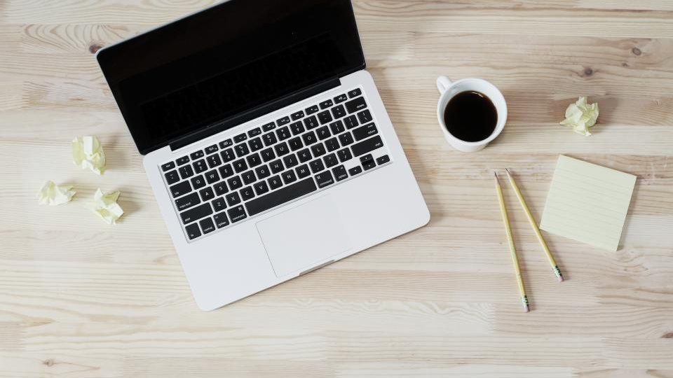 Un portatile chiuso su una scrivania di legno chiaro, con una tazza di caffè e due matite