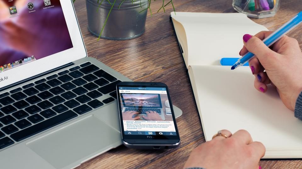 Una mano scrive un elenco su un taccuino aperto, accanto a un portatile e a uno smartphone