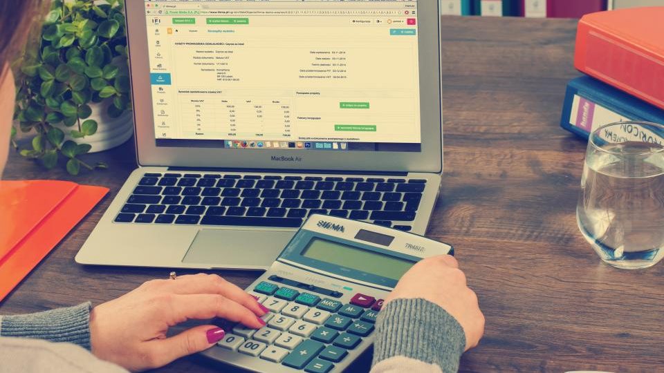 Un portatile con un foglio di calcolo aperto e due mani che usano una calcolatrice sulla scrivania